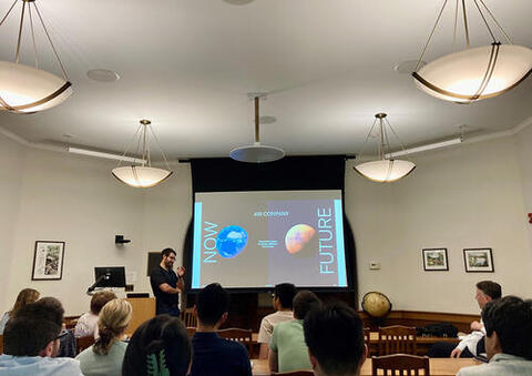 Dr. Staff Sheehan presents in front of a screen with images of the earth and Mars nexts to the words "Now" and "Future".